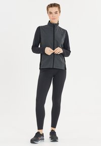 Schwarze Sportweste mit Reißverschluss, graues Langarmshirt darunter und schwarze Leggings. Getragen mit schwarzen Sportschuhen, die eine strukturierte Oberfläche haben.