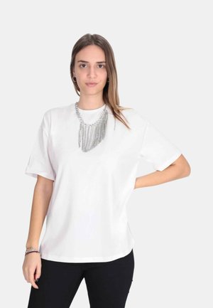 Donna che indossa una maglietta bianca semplice, una collana argentata con frange, pantaloni neri e braccialetti, in piedi con una mano sui fianchi.