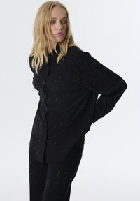 Camicia nera con bottoni, realizzata in un tessuto testurizzato, con piccoli dettagli lucidi. Maniche lunghe e vestibilità rilassata, abbinata a pantaloni neri.