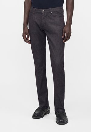 Άνδρας που φοράει σκούρα γκρι τζιν slim-fit και μαύρα δερμάτινα επίσημα παπούτσια στέκεται μπροστά σε απλό φόντο.