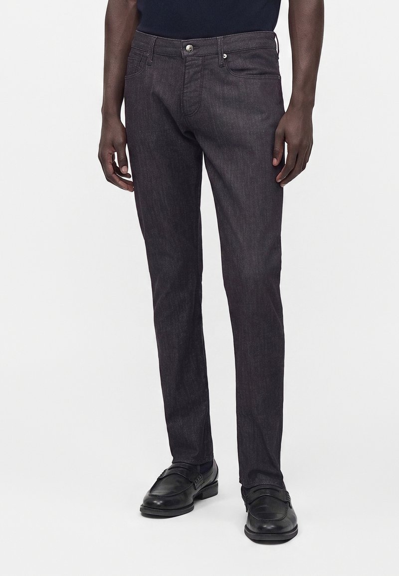 Άνδρας που φοράει σκούρα γκρι τζιν slim-fit και μαύρα δερμάτινα επίσημα παπούτσια στέκεται μπροστά σε απλό φόντο.