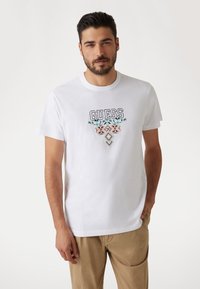 Weißes Baumwoll-T-Shirt mit schwarzem "GUESS"-Logo und mehrfarbiger geometrischer Stickerei darunter. Kurzärmlig, Rundhalsausschnitt und glatte Textur.
