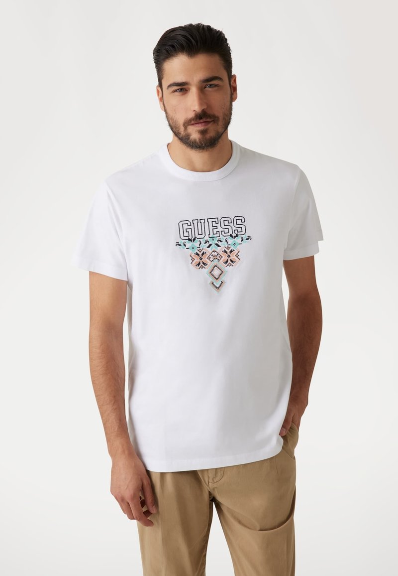 Weißes Baumwoll-T-Shirt mit schwarzem "GUESS"-Logo und mehrfarbiger geometrischer Stickerei darunter. Kurzärmlig, Rundhalsausschnitt und glatte Textur.