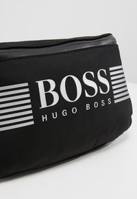 Sac en tissu noir avec les logos "BOSS" et "HUGO BOSS" imprimés en blanc. Présente des rayures horizontales de largeurs variées en accents.