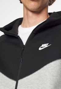 Nike zip-hoodie med en sort øverste sektion og lys grå nederste sektion, lavet af blødt stof med et fremtrædende logo på brystet.