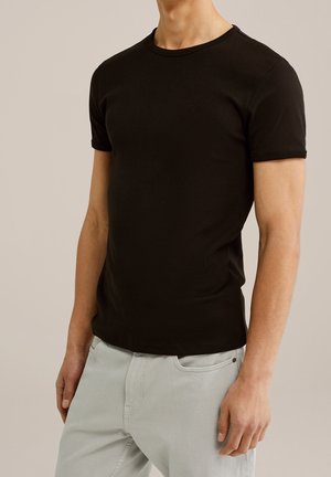 Homme portant un t-shirt noir uni à manches courtes et un pantalon gris clair, debout devant un fond neutre.