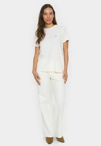 Camiseta de algodón color crema con un pequeño gráfico de lazo aqua en el pecho, combinada con pantalones de pierna ancha blancos y zapatos marrones de punta.