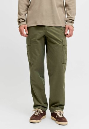 Personne portant un pantalon cargo vert olive, une chemise beige à manches longues et des baskets marron avec des lacets crème, debout devant un fond uni.
