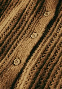 Neulehuppari lämpimissä ruskeissa sävyissä, jossa on teksturoitu kuvio, pystysuorat raidat ja neljä vaaleaa nappia.