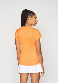 Femeie cu părul împletit, purtând o cămașă portocalie cu mâneci scurte și o fustă albă, văzută din spate pe un fundal deschis.