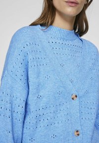 Svijetloplava pletena kardigan jakna s v-izrezom, koja ima teksturirani uzorak i tri dugmeta sprijeda.