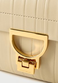Μπεζ τσάντα από δερμάτινο κέντημα με μεταλλικό κλείσιμο σε χρυσόχρωμο τόνο. Διαθέτει χαρακτηριστική κυκλική λαβή και λογότυπο στο κλείσιμο.