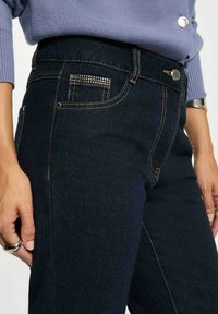 Jeans in denim scuro con vita alta, cuciture a contrasto e dettagli decorativi con borchie nella tasca. Texture liscia e silhouette aderente.