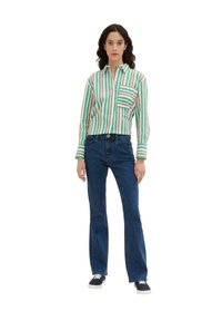 Gestreiftes, grün-weißes Hemd mit Kragen und aufgesetzter Tasche, kombiniert mit dunkelblauen Schlagjeans und marineblauen Sneakers. Weißer Hintergrund.