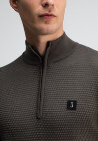 Grijze, gestructureerde gebreide pullover met een hoge kraag, een kwart-rits aan de voorkant en een zwart vierkant logo op de borst.