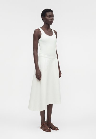 Γυναίκα στέκεται φορώντας λευκό αμάνικο ριμπ κοντόπουλο, ασύμμετρη λευκή φούστα και καφέ επίπεδα σανδάλια μπροστά σε μονόχρωμο λευκό φόντο.