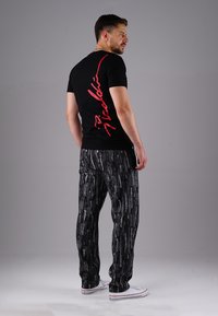 Schwarzes T-Shirt mit rotem Grafiktext auf dem Rücken, kombiniert mit gemusterten grau-schwarzen Hosen. Das Modell trägt weiße Schuhe und steht vor einem neutralen Hintergrund.