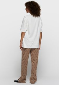 Camiseta blanca de algodón con mangas cortas, combinada con pantalones marrones con un patrón de logotipos repetidos. Modelo visto desde atrás.