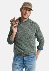 Grau gesprenkelter Strickpullover, darunter ein bluses Jeanshemd, blaue Jeans und eine beige Mütze mit Logo. Roter karierten Schal über die Schulter gelegt.