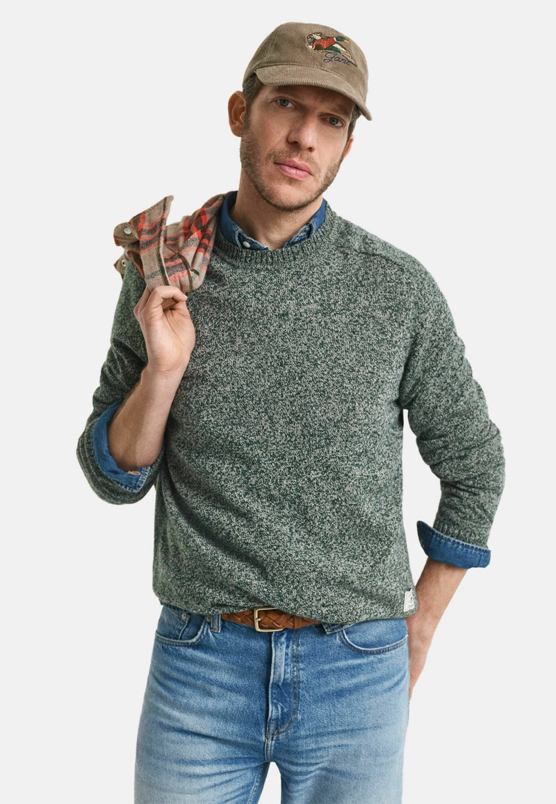 Grau gesprenkelter Strickpullover, darunter ein bluses Jeanshemd, blaue Jeans und eine beige Mütze mit Logo. Roter karierten Schal über die Schulter gelegt.