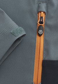 Grått tyg med en orange dragkedja, med en svart dragflik och ett kardborreband. Inkluderar kontrasterande sömmar och funktionella designinslag.