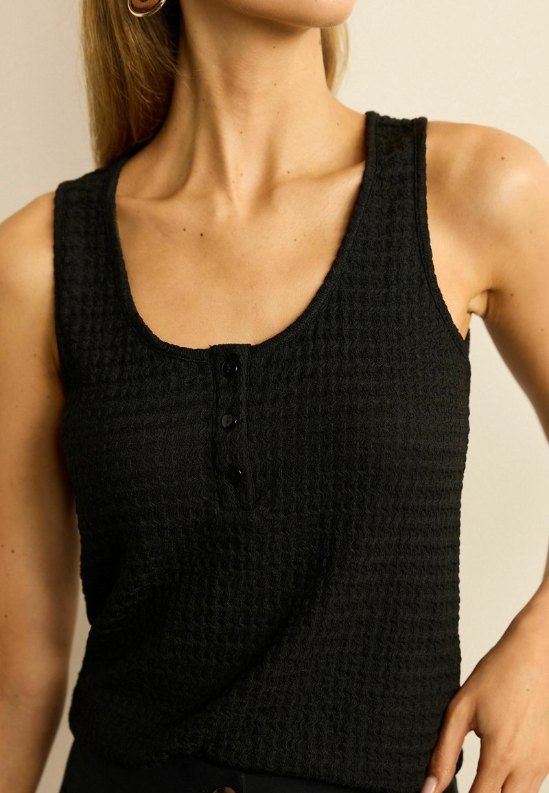 Black tank top with a textured waffle pattern, scoop neckline, and three buttons at the front. Material appears soft and lightweight.