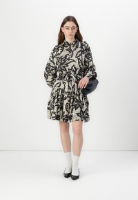 Abito floreale in bianco e nero con vita regolabile, maniche lunghe e gonna a balze. Modella abbinato a una borsa nera e ballerine nere.