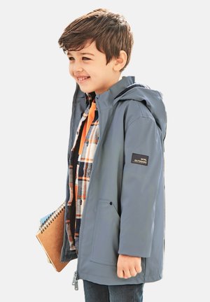 Giacca impermeabile grigio chiaro con cappuccio, dotata di un sottile patch con logo, abbinata a una camicia di flanella arancione a quadri.