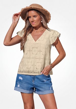 Vrouw die een beige blouse met korte mouwen en patroon draagt, blauwe opgerolde denimshorts, een strohoed en een armband met schelpenthema, poseren met hand op hoed.