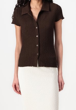 Femme portant une chemise à manches courtes marron foncé texturée avec des boutons et une jupe blanche texturée taille haute, debout devant un fond blanc.