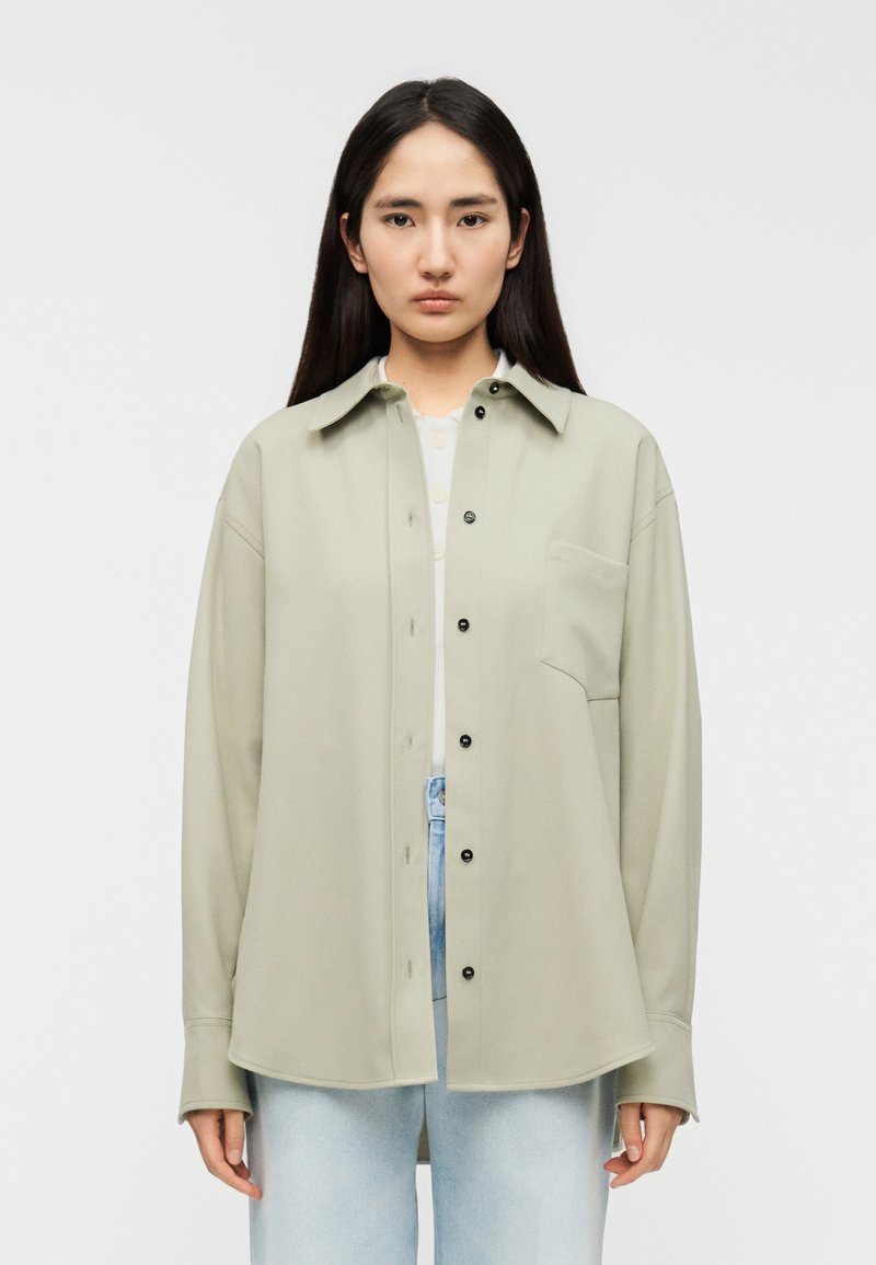 Υπερμεγέθη πουκάμισο σε ελιά πράσινο χρώμα, κατασκευασμένο από απαλό ύφασμα, με τσέπη στο στήθος και μαύρα κουμπιά, φορεμένο πάνω από λευκό τοπ.