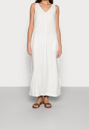 Woman wearing a sleeveless white maxi dress with a V-neck, standing with arms at sides, and brown ankle-strap flat sandals.