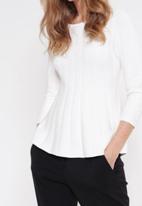 Haut blanc à côtes avec finition texturée, encolure ronde et manches trois-quarts, associé à un pantalon noir, créant une silhouette décontractée.