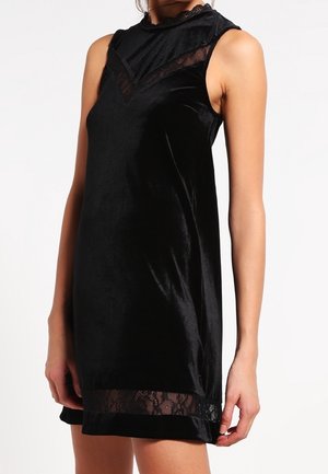 Czarna aksamitna sukienka bez rękawów z koronkową lamówką przy wysokim dekolcie, górnej części klatki piersiowej i na dole, noszona przez stojącą osobę na białym tle.