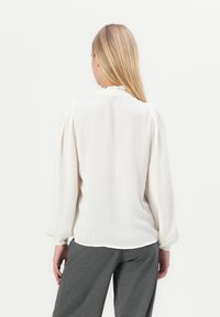 En blond kvinde iført en hvid bluse med lange ærmer og rynket krave samt grå bukser, står med ryggen til mod en hvid baggrund.