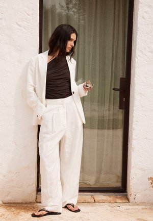 Femeie îmbrăcată într-un costum alb cu pantaloni și top negru, ținând o ceașcă de sticlă, stând lângă o ușă de sticlă cu draperii, într-un decor rustic în aer liber.