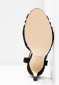 Sandale à talon haut en daim noir avec fines lanières, semelle beige, taille 37, positionnée debout contre un mur d'angle blanc.