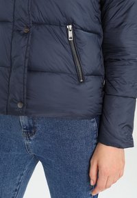 Marineblå dunjakke med quiltet design, sølv lynlåslomme og trykknapper. Skinnende stof med en glat tekstur. Båret med blå jeans.