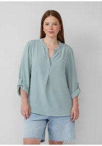 Helles tealfarbendes Blusenshirt mit einem kragenlosen Design, Drei-Viertel-Ärmeln mit Aufroll-Tab, und einer dezenten gestreiften Textur, kombiniert mit Jeansshorts.