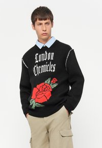 Jonge man in een zwarte trui met de tekst "London Chronicles" en een rode rozenafbeelding, beige cargobroek en een blauw gestreept overhemd met kraag.