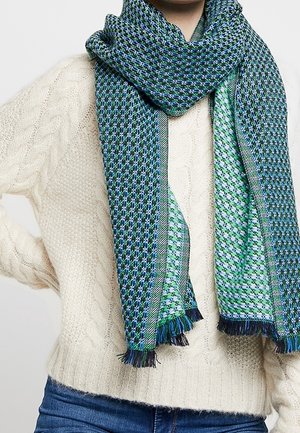 Écharpe tricotée aux motifs bleus, verts et noirs enroulée autour du cou, portée sur un pull en tricot torsadé crème et un jean bleu.