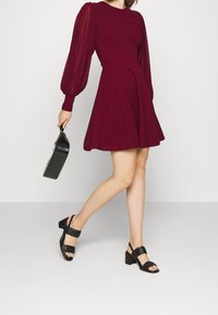 Kvinna i en vinröd klänning med genomskinliga puffärmar, svarta klacksandaler, som håller en svart texturerad handväska, går mot en enkel bakgrund.