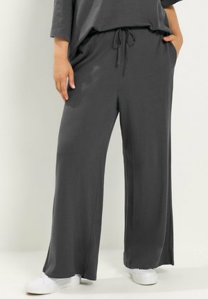 Person trägt lockere, dunkelgraue Hose mit Kordelzug in der Taille und weiße Sneaker, steht mit einer Hand in der Tasche vor neutralem Hintergrund.