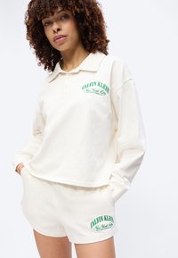 Γυναίκα με σγουρά μαλλιά, φοράει εκρού πόλο με μακριά μανίκια και αντίστοιχα σορτς με πράσινο κείμενο «Calvin Klein New York City».