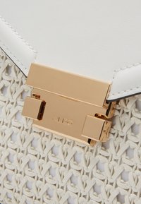 Bílá kabelka s tkanou texturou a hladkým povrchem, zdobená zlatým zapínáním a vygravírovaným logem.