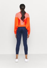 Röd och orange färgblockerad jacka med ribbat mönster, kombinerad med marinblå leggings med vita ränder på sidorna. Vita sneakers.