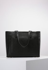 Sac fourre-tout en cuir noir avec une surface texturée, deux poignées robustes et une poche extérieure. Présente une forme structurée et des accents dorés.