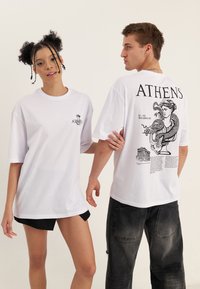 Jonge vrouw en man dragen witte oversized T-shirts met "ATHEN" en oude Griekse afbeeldingen, staand tegen een effen achtergrond.