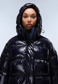 Giacca a vento imbottita in materiale blu navy lucido, con colletto alto e cappuccio, pannelli cuciti e chiusura frontale con zip per l'isolamento.