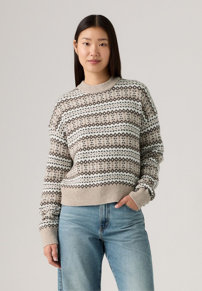 Gebreide trui in beige met patroonbanden van bruine en witte diamanten, ronde halslijn, geribde manchetten en zoom. Gecombineerd met lichtblauwe jeans.